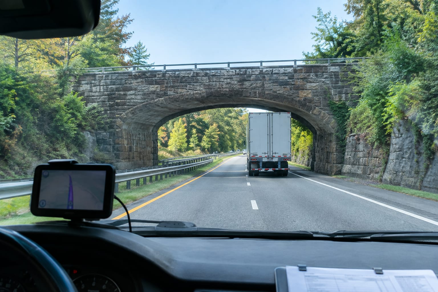 Driver's view of a moving truck approaching a low stone-arch overpass marked with a yellow clearance-height sign