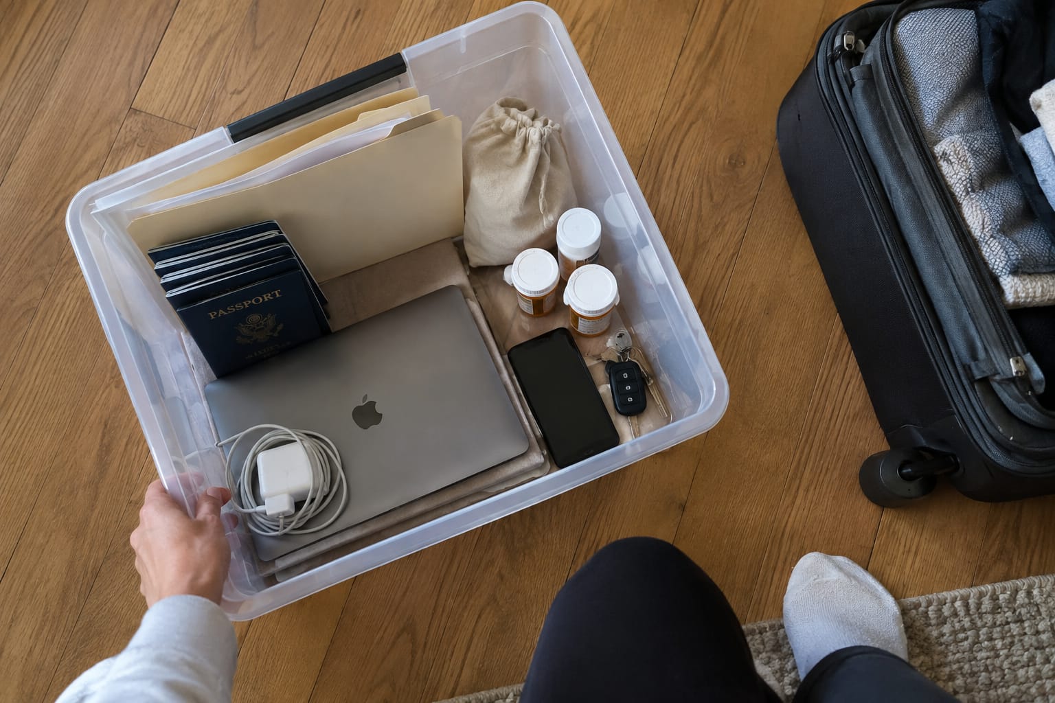 Overhead view of a clear bin with passports, jewelry, medication, and keys labeled 'GOES WITH ME' beside a suitcase.