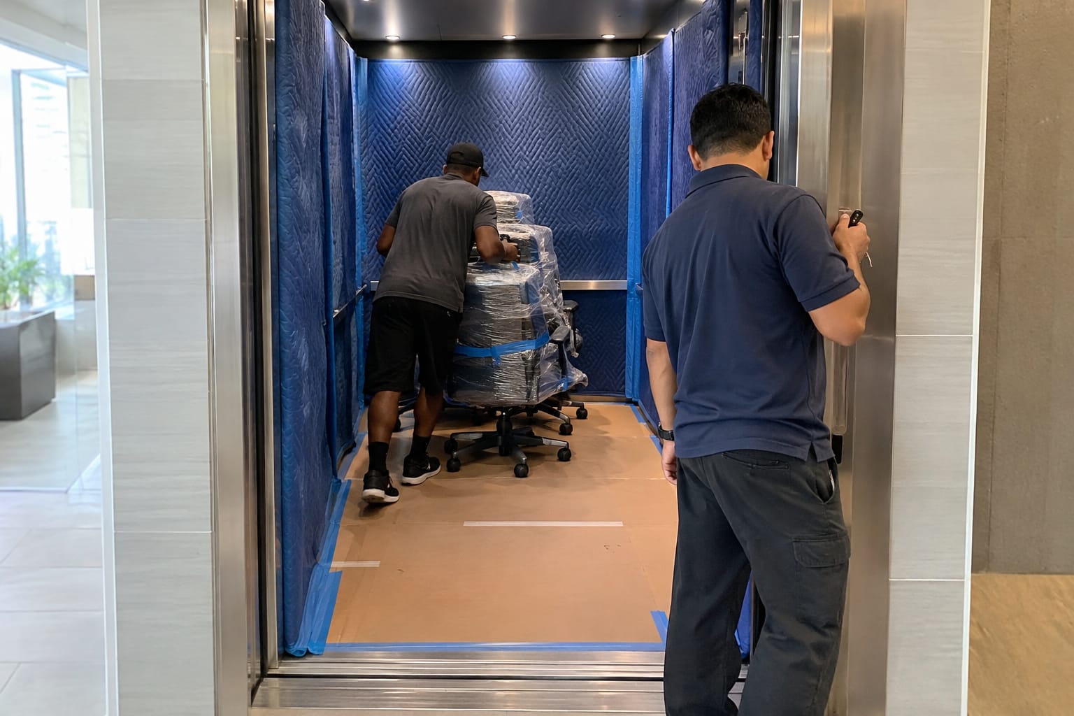 Padded service elevator with corner guards and a reserved sign as movers roll in wrapped office chairs.