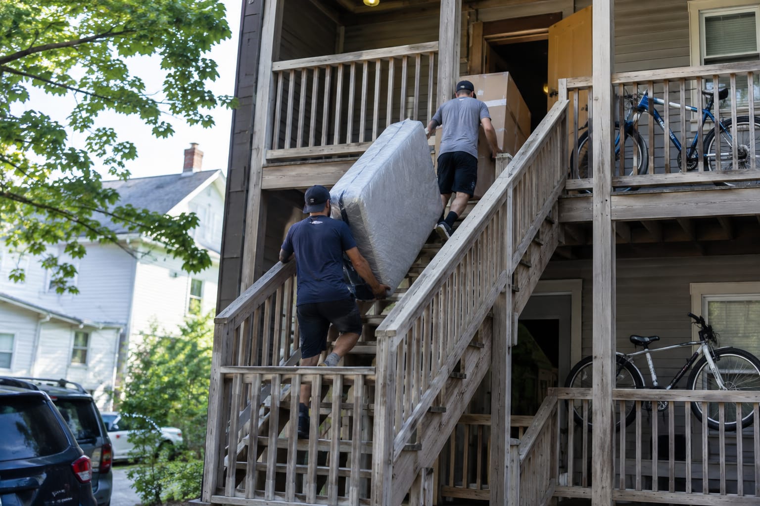 Two movers carrying a mattress and wardrobe box up an exterior triple-decker staircase to a propped door