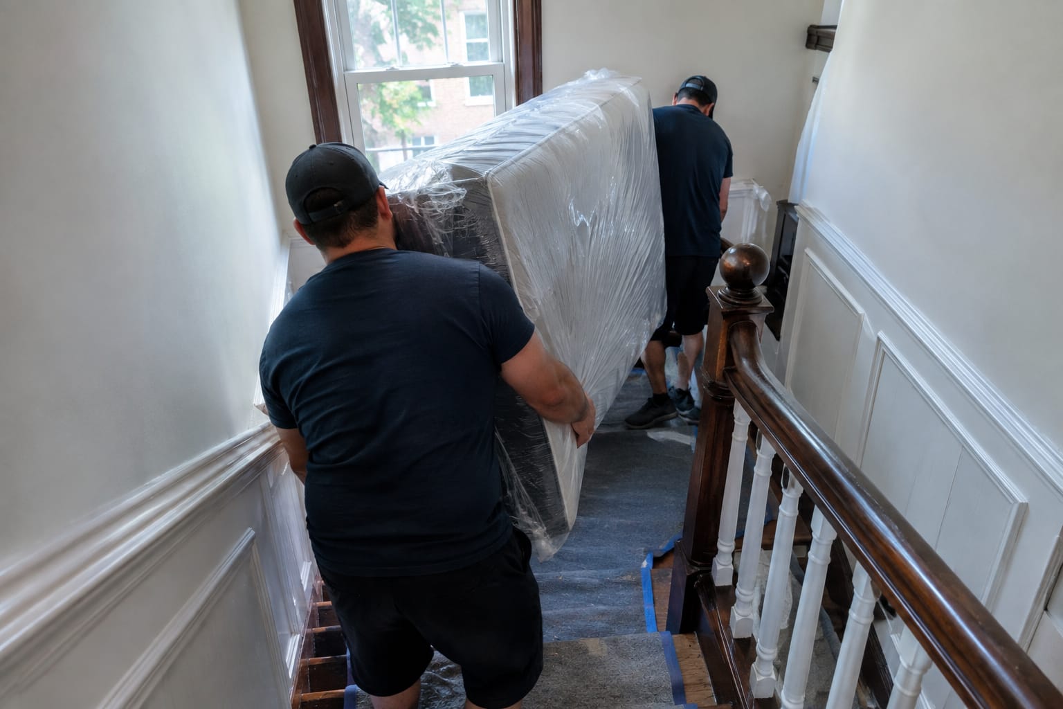 Two movers turning a shrink-wrapped mattress around a sharp narrow staircase landing with a floor runner on the treads.