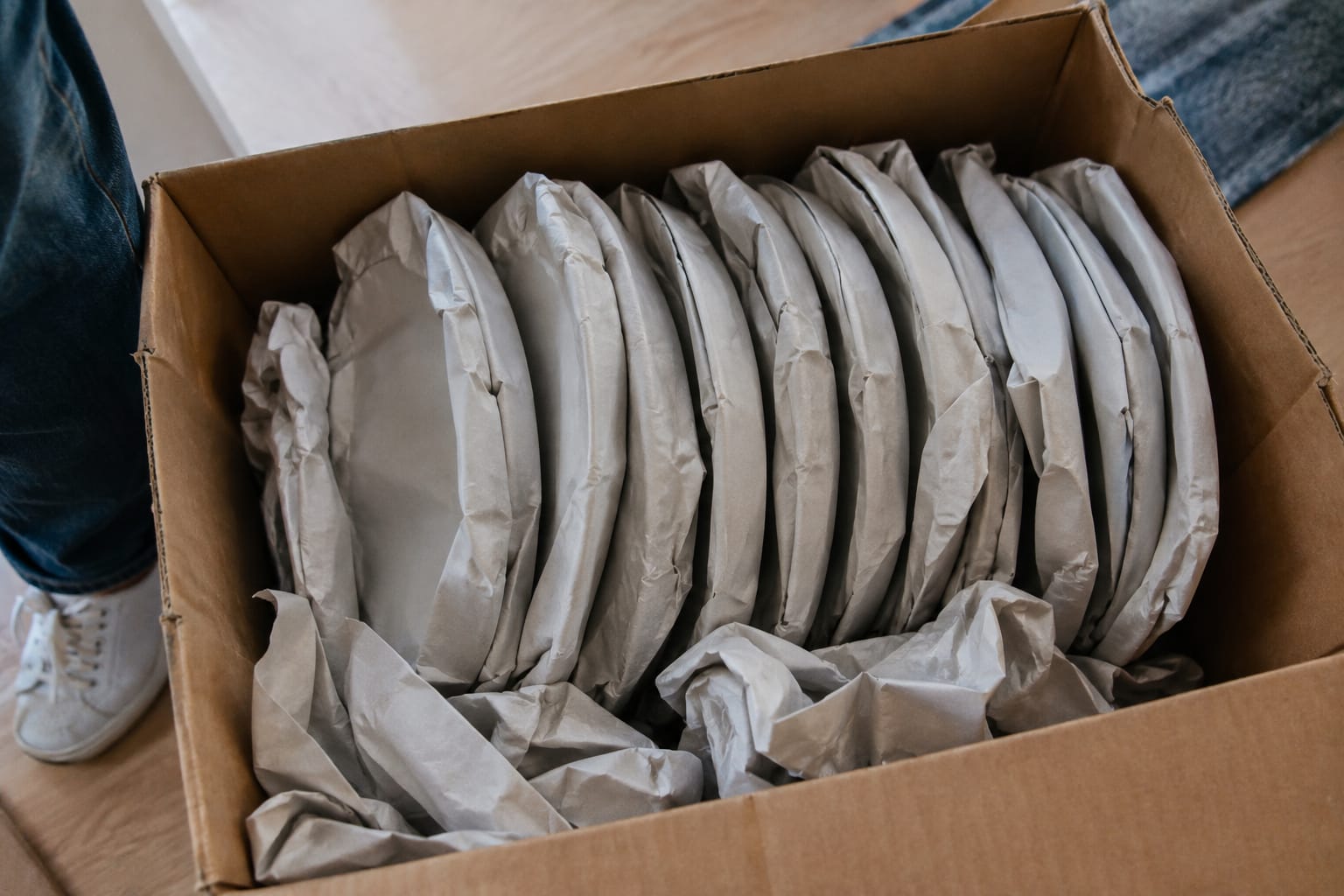 Hands wrapping a dinner plate in packing paper above a box of wrapped plates standing vertically