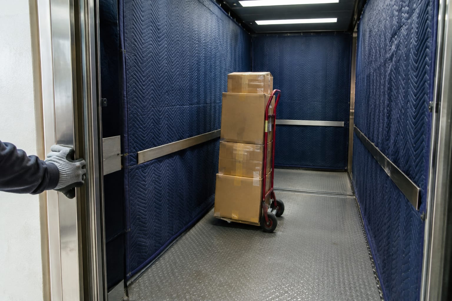 Mover pushing a dolly stacked with boxes toward an open, padded freight elevator at a high-rise loading dock