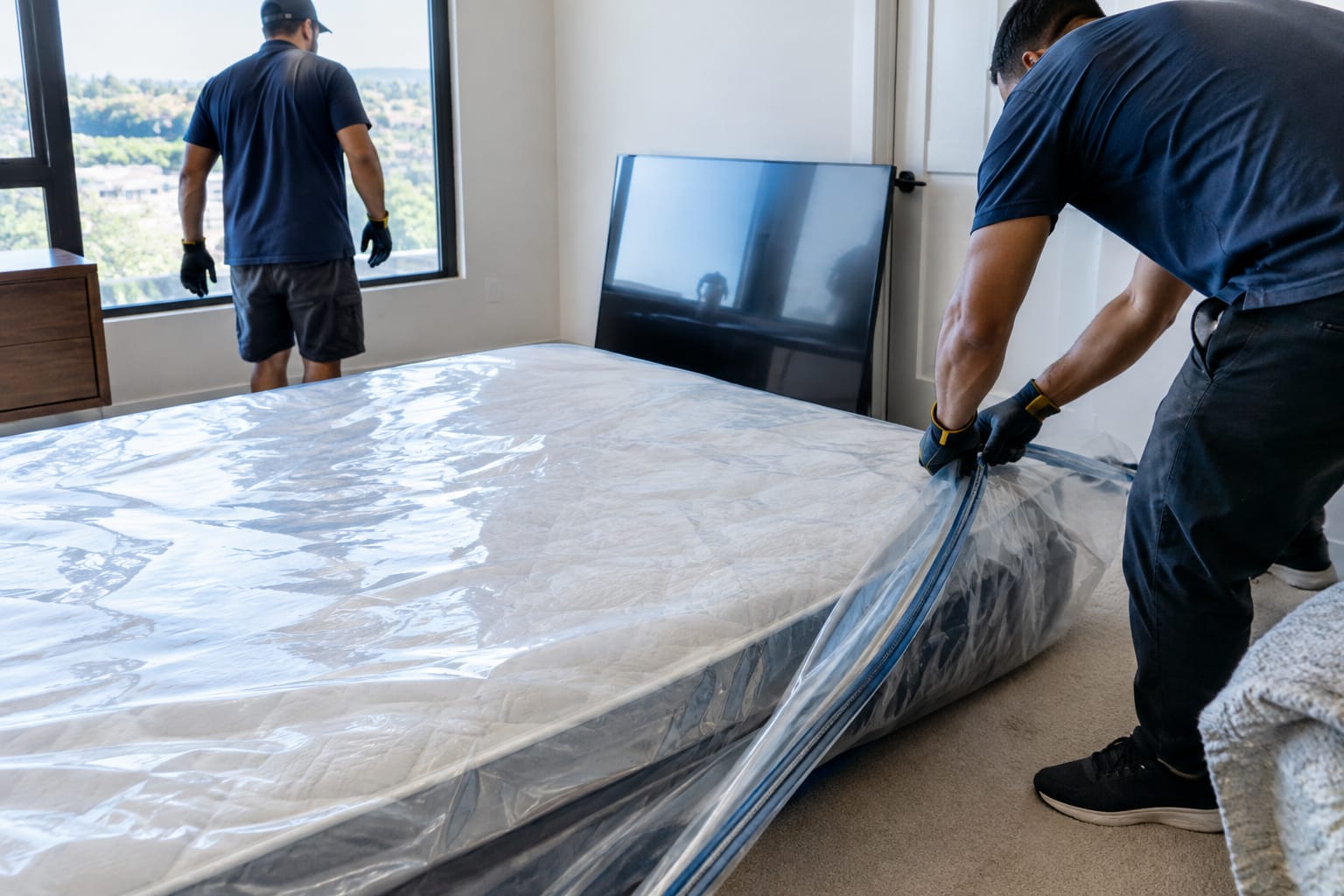 Queen mattress in a clear plastic bag beside an upright flat-screen TV box in an empty bedroom
