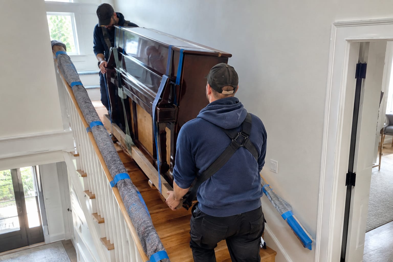 Two movers easing an upright piano down interior stairs on a piano board with padded banister edges