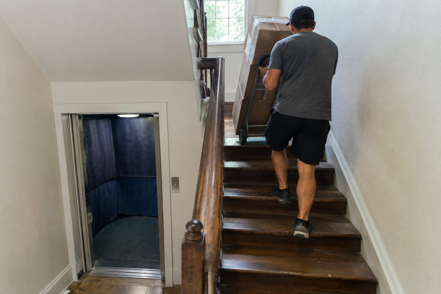 Two movers using shoulder lifting straps to carry a dresser up a narrow wooden staircase